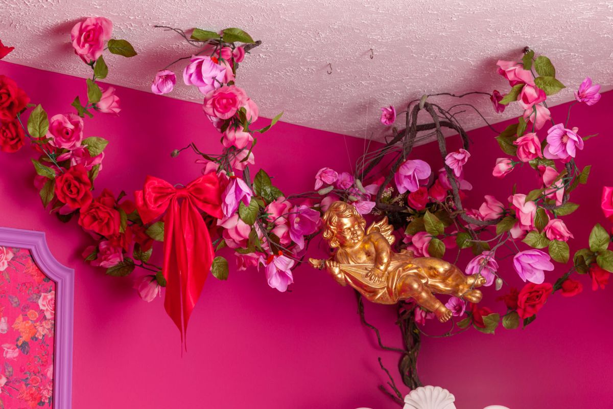 A golden cherub hangs among artificial flowers and greenery on a pink wall.